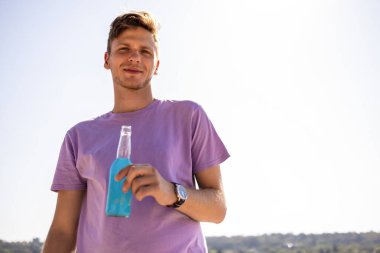 Genç yakışıklı adam sahilde mavi içki içiyor..