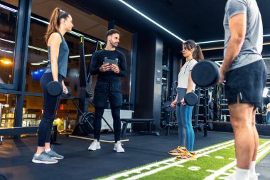 Bir grup insan halter ağırlıklarıyla grup antrenmanına giderken kişisel antrenörleri seti açıklayıp sayıyor..