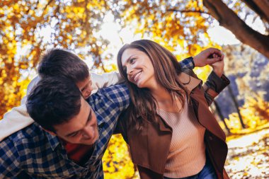 Gülümseyen baba, sonbahar parkında küçük oğlunu sırtında taşır. Annem gülümsüyor. Parktaki aile.