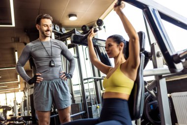 Genç, formda bir kadın spor salonundaki bir makinede omuz ve kollarını çalıştırırken özel antrenörü ona yardım ediyor..