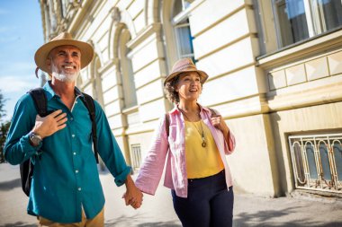 Yeni şehri keşfeden yaşlı evli bir çift. El ele tutuşuyorlar ve şapka ve sırt çantası takıyorlar..