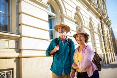 Yeni şehri keşfeden yaşlı evli bir çift. El ele tutuşuyorlar ve şapka ve sırt çantası takıyorlar..