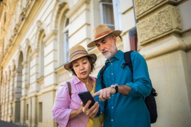 Yaşlı evli çift yeni şehri keşfediyor. Navigasyon ve harita için telefonlarını kullanıyorlar. Şapka ve sırt çantası takıyorlar..