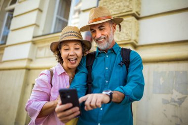 Yaşlı evli çift yeni şehri keşfediyor. Navigasyon ve harita için telefonlarını kullanıyorlar. Şapka ve sırt çantası takıyorlar..
