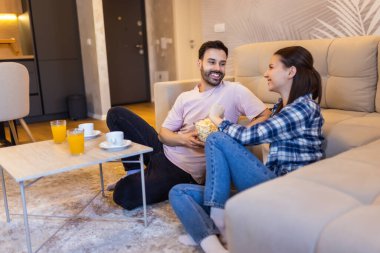 Oturma odasındaki kanepede dinlenen bir çift. Patlamış mısır yiyip meyve suyu içiyorlar..