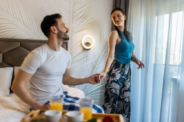 Pijamalı bir çift. Kız arkadaşı pencereden bakıyor ve erkek arkadaşı sabah hala yataktayken onun elini tutuyor..