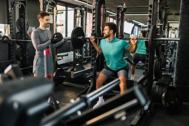 Formda olan adam kişisel antrenörünün yardımıyla halterle çömeliyor..