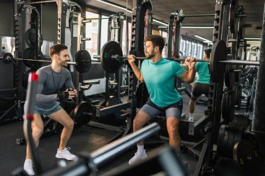 Formda olan adam kişisel antrenörünün yardımıyla halterle çömeliyor..