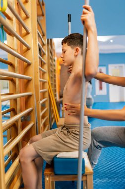 Küçük çocuğun terapi amaçlı kinezi terapisi. Bir çocuk belkemiğini ve leğen kemiğini düzleştirmek için egzersiz yaparken tabureye tutunuyor..