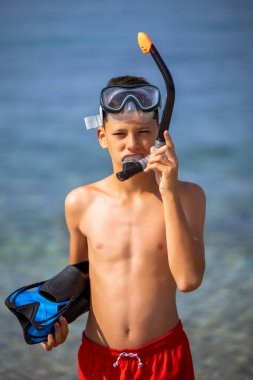 Dalış maskesi takan çocuk, şnorkel ve yüzgeçler suya dalmaya hazırlanıyor. Kumsalda güneşli bir gün.