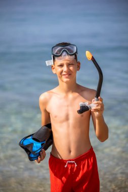 Dalış maskesi takan çocuk, şnorkel ve yüzgeçler suya dalmaya hazırlanıyor. Kumsalda güneşli bir gün.