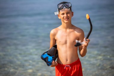 Dalış maskesi takan çocuk, şnorkel ve yüzgeçler suya dalmaya hazırlanıyor. Kumsalda güneşli bir gün.