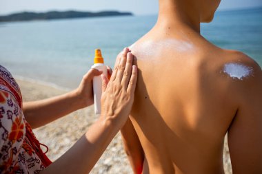 Anne oğlunun arkasına güneş kremi sürüyor. Kumsalda güneşli sıcak bir gün.