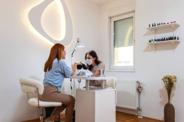 Güzellik salonundaki kadın manikürle tırnaklarını yaptırıyor. Maske ve eldiven takıyor..