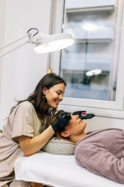 Bir kadının kaşlarına fırça kullanarak kaş boyası süren bir güzellik uzmanı..