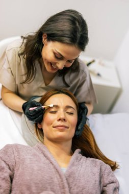 Bir kadının kaşlarına fırça kullanarak kaş boyası süren bir güzellik uzmanı..