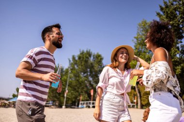 Canlı bir plaj partisinde kokteyl içen bir grup arkadaş. Arka planda müzik, renkli içecekler ve okyanusun üzerinde güneş batarken sahne enerji, eğlence ve yaz kutlamalarıyla dolu. Saf sahil titreşimleri ve gençlik sevinci.
