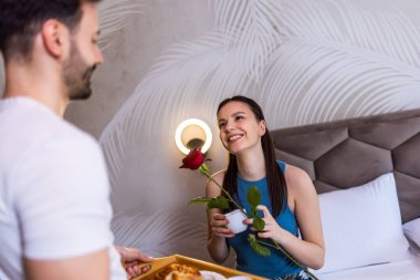 Erkek arkadaşı kız arkadaşı için kahvaltıyı yatağa getirdi. Kız arkadaşı erkek arkadaşından aldığı gülü kokluyor. Tepside kruvasan, portakal suyu ve kahve var..