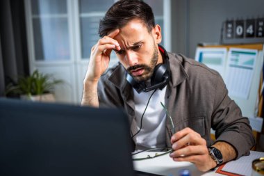 Evde dizüstü bilgisayarıyla çalışan yorgun genç bir adam. Kulaklıkları boynunda.. 