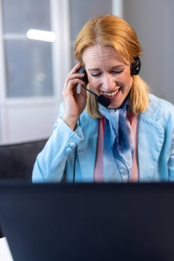 Kulaklık ve mikrofon takan bir kadın dizüstü bilgisayar kullanırken işine odaklanmış. Görüntü, profesyonel bir ofis ortamında uzaktan çalışma, dijital iletişim ve sanal toplantıları temsil etmektedir..