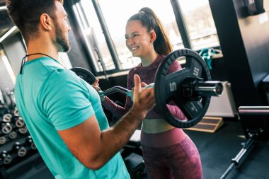 Genç bir kadın spor salonunda ağırlıklarla omuz presi yaparken antrenörü onu fark ediyor..