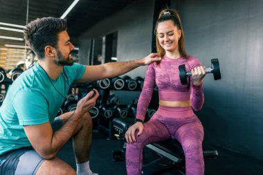 Destekleyici genç bir adam, modern bir spor salonunda ağırlık kaldıran bir kadına yardım ediyor..