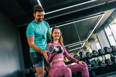Destekleyici genç bir adam, modern bir spor salonunda ağırlık kaldıran bir kadına yardım ediyor..