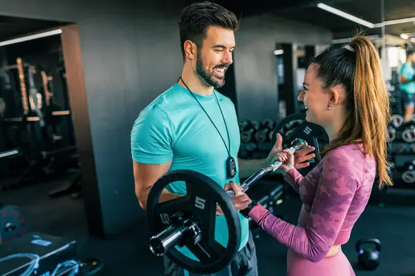 Genç bir kadın spor salonunda ağırlıklarla omuz presi yaparken antrenörü onu fark ediyor..