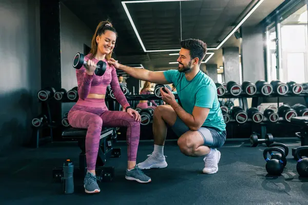 Destekleyici genç bir adam, modern bir spor salonunda ağırlık kaldıran bir kadına yardım ediyor..