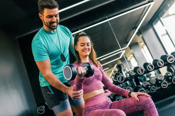 Destekleyici genç bir adam, modern bir spor salonunda ağırlık kaldıran bir kadına yardım ediyor..