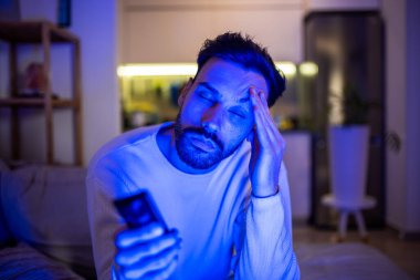 Uykulu genç bir adam loş bir odada televizyon izler, gözleri yorgunluktan ağırlaşır.. 