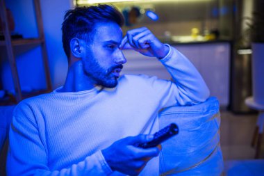 Uykulu genç bir adam loş bir odada televizyon izler, gözleri yorgunluktan ağırlaşır.. 