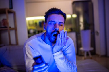 Uykulu genç bir adam loş bir odada televizyon izler, gözleri yorgunluktan ağırlaşır.. 