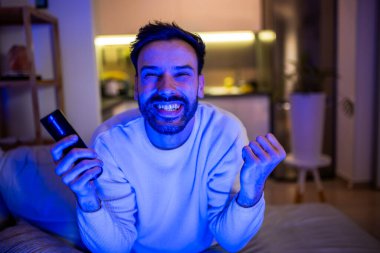 Karanlık bir odada televizyon izleyen neşeli bir adam. Yüzü ekranın yumuşak parıltısıyla aydınlanıyor.. 