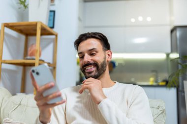 Genç bir adam oturma odasındaki koltukta rahat bir şekilde oturuyor, akıllı telefonunu kullanıyor..