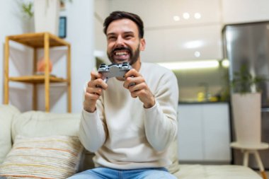 Oturma odasındaki kanepede oturan genç bir adam elinde kumandayla video oyunları oynamaya odaklanmış..