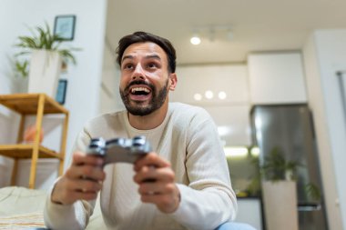 Oturma odasındaki kanepede oturan genç bir adam elinde kumandayla video oyunları oynamaya odaklanmış..
