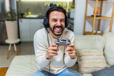 Genç bir adam oturma odasında bir kanepede oturuyor, kulaklık takıyor ve video oyunları oynarken bir oyun kumandası tutuyor.. 