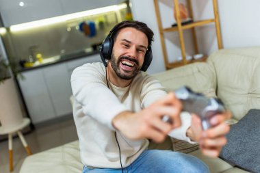 Genç bir adam oturma odasında bir kanepede oturuyor, kulaklık takıyor ve video oyunları oynarken bir oyun kumandası tutuyor.. 