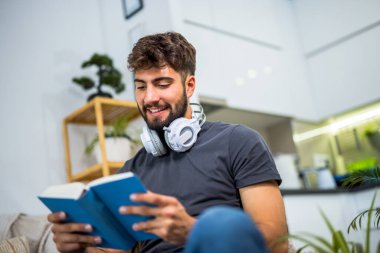 Genç, sakallı bir adam rahat bir şekilde bir kanepede oturur rahat bir oturma odasında, tamamen bir kitaba gömülmüş. Evinin sessizliğiyle çevrili, huzurlu bir yalnızlık anının tadını çıkarıyor, sayfalarla ekran zamanını değiş tokuş ediyor..