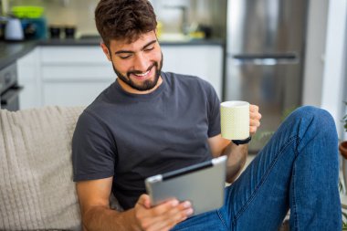 Genç sakallı bir adam, rahat bir oturma odasında kanepede oturuyor, tabletine dalmış. Dijital rahatlama anının tadını çıkarıyor.. 
