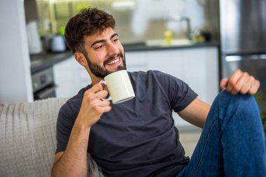 Genç sakallı bir adam sıcak oturma odasındaki kanepede rahatça oturup sıcak bir kahvenin tadını çıkarıyor. Basit ama huzurlu bir rahatlama anı. Evin sakin atmosferi, yavaş ve yansıtıcı bir mola için mükemmel bir ortam sağlıyor.. 