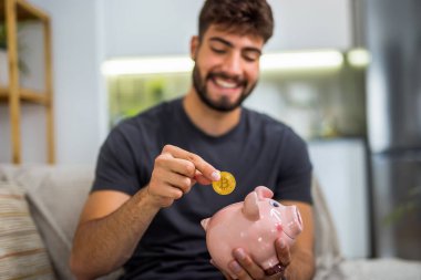 Genç bir adam geleneksel bir kumbara içine bir bitcoin sembolü ya da temsili yerleştirir, modern kripto para birimini eski moda tasarruf yöntemleriyle birleştirir.. 