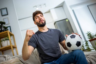 Oturma odasında oturan genç bir adam, bir futbol maçına dalmış, en sevdiği takım oynarken heyecanla tezahürat yapıyor. Elinde bir futbol topu tutarak spor ve takım ruhuna olan tutkusunu ifade ediyor..