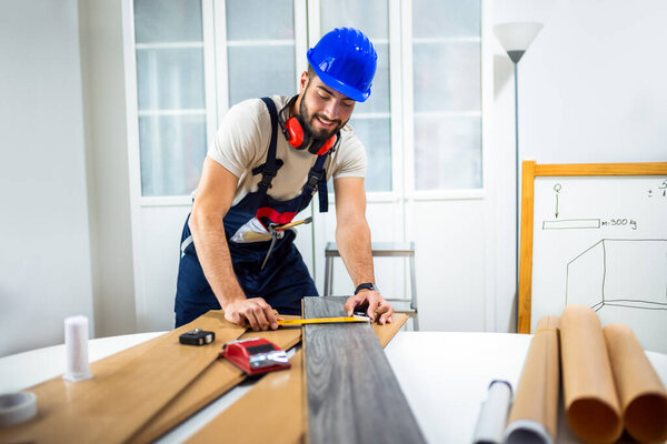 A young man works diligently on renovating his apartment, focusing on the installation of laminate flooring. With the right tools in hand, he carefully measures and fits the laminate panels into place, transforming the space with his DIY skills. 