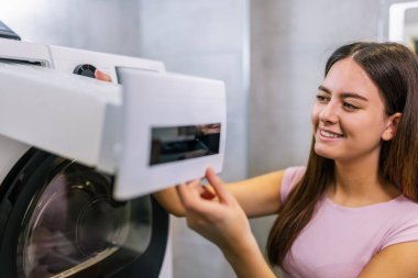 Genç bir kız çamaşır kurutma makinesinden su kabını çıkarıyor. Makinenin iyi çalışması için düzenli bakım yapıyor..