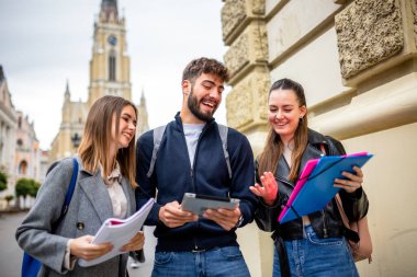 Üç ergen öğrenciden oluşan bir grup birlikte ders çalışıyorlar. Ellerinde kitap ve tablet var. Eğitimsel içeriği tartışıyor ve paylaşıyorlar..