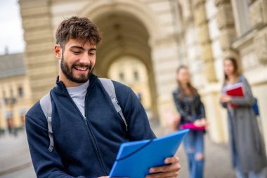 Kendinden emin bir öğrenci ön planda duruyor, elinde bir klasör tutarken kameraya gülümsüyor. Arkasından iki öğrenci daha görülüyor, biraz bulanık, bu da akademik ya da sosyal bir ortam olduğunu gösteriyor..