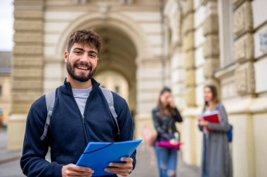 Kendinden emin bir öğrenci ön planda duruyor, elinde bir klasör tutarken kameraya gülümsüyor. Arkasından iki öğrenci daha görülüyor, biraz bulanık, bu da akademik ya da sosyal bir ortam olduğunu gösteriyor..