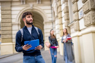 Kendinden emin bir öğrenci ön planda duruyor, elinde bir klasör tutarken kameraya gülümsüyor. Arkasından iki öğrenci daha görülüyor, biraz bulanık, bu da akademik ya da sosyal bir ortam olduğunu gösteriyor..